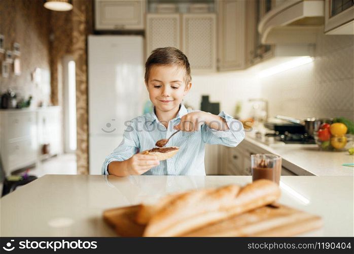 Young boy smears melted chocolate on bread. Cute male kid cooking on the kitchen. Happy child prepares and tastes sweet dessert at the counter