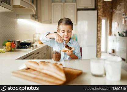Young boy smears melted chocolate on bread. Cute male kid cooking on the kitchen. Happy child prepares and tastes sweet dessert at the counter. Young boy smears melted chocolate on bread