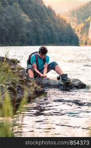 Young boy takes pure water from a river and holds it in the hands. He is sitting on a rock over the river, rests during a hike, spends a vacation on wandering with backpack, he is wearing sport summer clothes