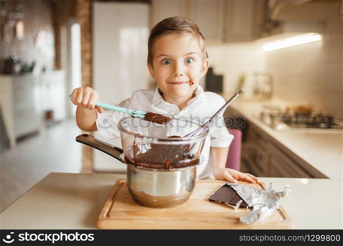 Young boy tastes melted chocolate in a bowl. Kid cooking on the kitchen. Happy child prepares sweet dessert. Young boy tastes melted chocolate in a bowl