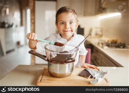 Young boy tastes melted chocolate in a bowl. Kid cooking on the kitchen. Happy child prepares sweet dessert. Young boy tastes melted chocolate in a bowl