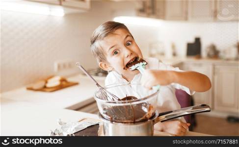 Young boy tastes melted chocolate in a bowl. Kid cooking on the kitchen. Happy child prepares sweet dessert at the counter