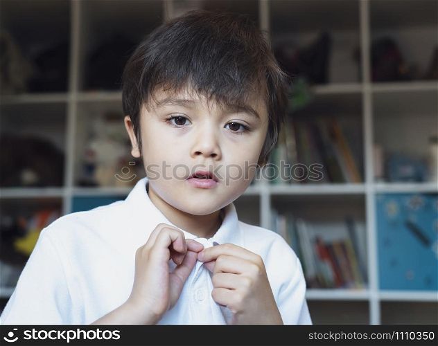 Young boy trying to close white polo shirt buttons, Emotional portrait of Kid sitting in bed looking out deep in through while tried to button up his school shirt, Child get ready for school.