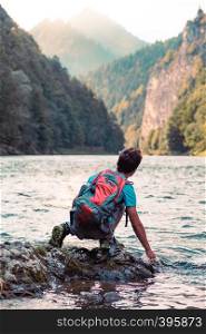 Young boy with backpack sitting on rock over a river, holding hand in water, looking at mountains peaks, he is wearing sports summer clothes