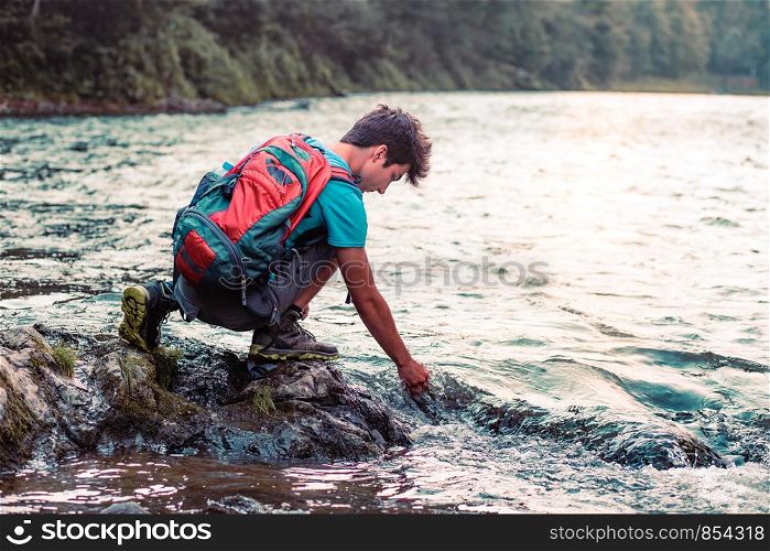 Young boy with backpack sitting on rock over a river, holding hand in water, looking down a water, he is wearing sports summer clothes