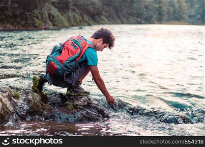 Young boy with backpack sitting on rock over a river, holding hand in water, looking down a water, he is wearing sports summer clothes