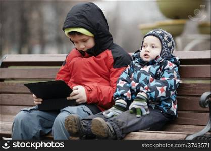 Young brothers sit together on a wooden park bench in warm winter clothing with the older child using a tablet computer