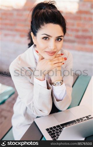 Young brunette businesswoman with charming eyes, gentle hands with red manicure wearing watch on hand and white coat holding hands under chin sitting near her laptop having rest after hard work