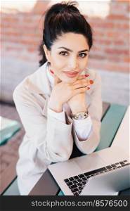 Young brunette businesswoman with charming eyes, gentle hands with red manicure wearing watch on hand and white coat holding hands under chin sitting near her laptop having rest after hard work
