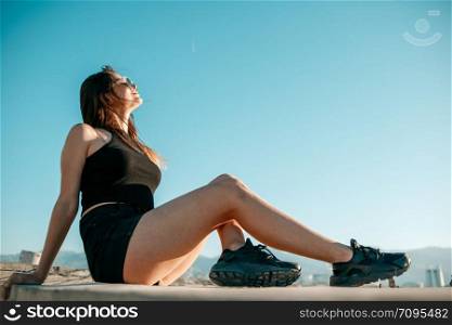 young brunette girl in black sneakers,black shorts and a black short top sitting on the roof of a tall building