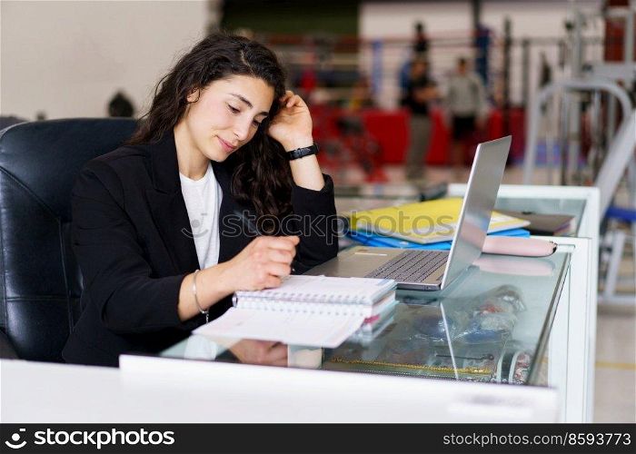 Young brunette in smart casual clothes touching hair and writing in notebook while sitting at desk with laptop during psychology session in gym. Sports psychologist making notes during work