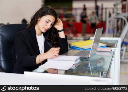 Young brunette in smart casual clothes touching hair and writing in notebook while sitting at desk with laptop during psychology session in gym. Sports psychologist making notes during work