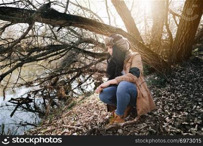 Young brunette woman crouched on the bank of a river wearing a jacket and a russian leather bomber hat with the ground full of autumn leaves