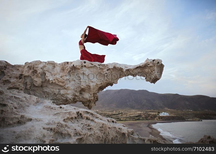 Young brunette woman dancing above a rock near sea coast wearing a red dress