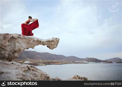 Young brunette woman dancing above a rock near sea coast wearing a red dress
