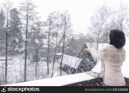 Young brunette woman, dressed casual, stands on a balcony with her hands stretched, reaching to catch the snowflakes, that fall over the houses.