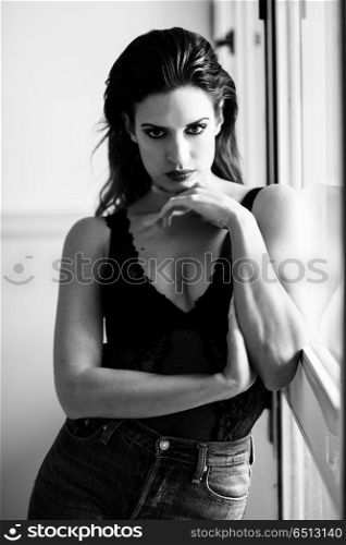Young brunette woman looking at camera near a window. . Young brunette woman looking at camera near a window. Attractive girl, model of fashion in lingerie and jeans. Black and white photograph.
