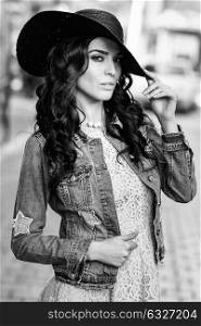 Young brunette woman, model of fashion, wearing denim jacket, hat, and dress. Pretty caucasian girl with long wavy hairstyle in urban background. Black and white.