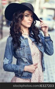 Young brunette woman, model of fashion, wearing denim jacket, hat, and pink dress. Pretty caucasian girl with long wavy hairstyle in urban background.