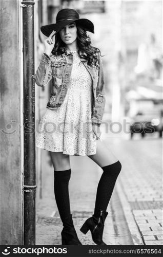 Young brunette woman, model of fashion, wearing denim jacket, hat, long socks and dress. Pretty caucasian girl with long wavy hairstyle in urban background.