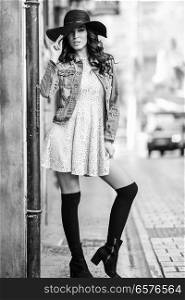 Young brunette woman, model of fashion, wearing denim jacket, hat, long socks and dress. Pretty caucasian girl with long wavy hairstyle in urban background.