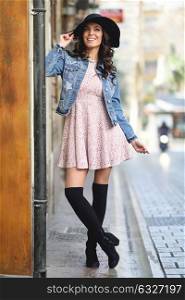 Young brunette woman, model of fashion, wearing denim jacket, hat, long socks and pink dress. Pretty caucasian girl with long wavy hairstyle smiling. Female in urban background.