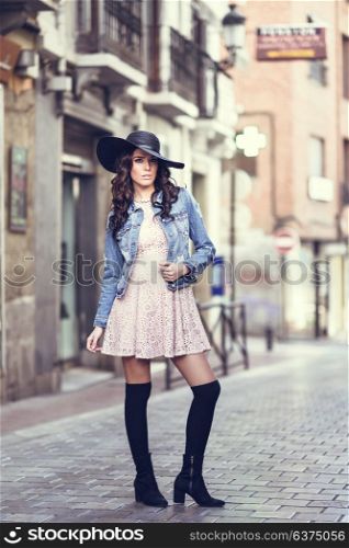 Young brunette woman, model of fashion, wearing denim jacket, hat, long socks and pink dress. Pretty caucasian girl with long wavy hairstyle in urban background.