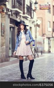 Young brunette woman, model of fashion, wearing denim jacket, hat, long socks and pink dress. Pretty caucasian girl with long wavy hairstyle in urban background.