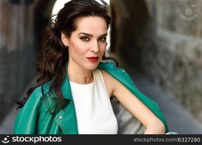 Young brunette woman, model of fashion, wearing green modern jacket. Pretty caucasian girl with long wavy hairstyle. Female with red lips in urban background.