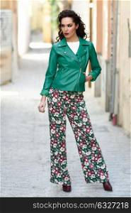 Young brunette woman, model of fashion, wearing green modern jacket and flower pants. Pretty caucasian girl with long wavy hairstyle. Female with red lips in urban background.