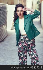 Young brunette woman, model of fashion, wearing green modern jacket and flower pants. Pretty caucasian girl with long wavy hairstyle. Female with red lips in urban background.