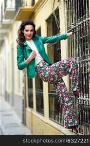 Young brunette woman, model of fashion, wearing green modern jacket and flower pants. Pretty caucasian girl with long wavy hairstyle smiling. Female with red lips in urban background.