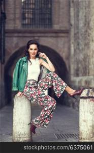 Young brunette woman, model of fashion, wearing green modern jacket and flower pants. Pretty caucasian girl with long wavy hairstyle smiling. Female with red lips in urban background.