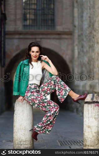 Young brunette woman, model of fashion, wearing green modern jacket and flower pants. Pretty caucasian girl with long wavy hairstyle smiling. Female with red lips in urban background.