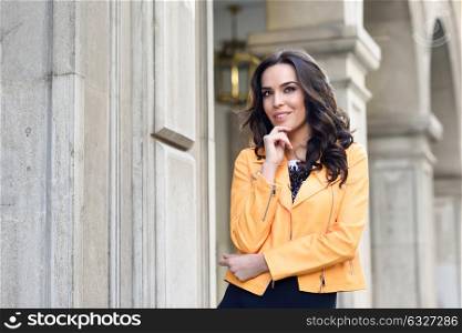 Young brunette woman, model of fashion, wearing orange modern jacket. Pretty caucasian girl with long wavy hairstyle. Thoughtful female in urban background.