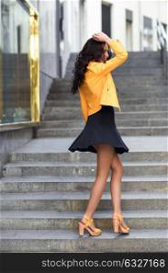 Young brunette woman, model of fashion, wearing orange modern jacket and blue skirt dancing in stairs. Pretty caucasian girl with long wavy hairstyle. Funny female in urban background.