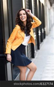 Young brunette woman, model of fashion, wearing orange modern jacket and blue skirt, laughing in urban background.