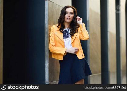 Young brunette woman, model of fashion, wearing orange modern jacket and blue skirt in urban background.