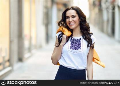 Young brunette woman, model of fashion, wearing orange modern jacket and blue skirt, smiling in urban background.