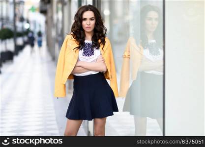 Young brunette woman, model of fashion, wearing orange modern jacket and blue skirt, standing in urban background.