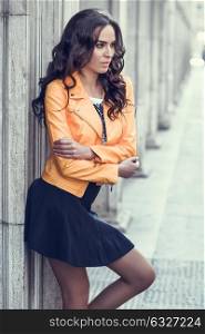 Young brunette woman, model of fashion, wearing orange modern jacket and blue skirt. Pretty caucasian girl with long wavy hairstyle. Female in urban background.