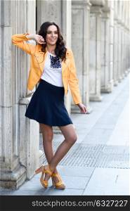 Young brunette woman, model of fashion, wearing orange modern jacket and blue skirt. Pretty caucasian girl with long wavy hairstyle smiling. Female raising her arms in urban background.
