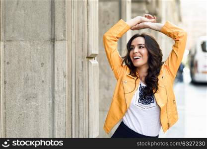 Young brunette woman, model of fashion, wearing orange modern jacket and blue skirt. Pretty caucasian girl with long wavy hairstyle smiling. Female raising her arms in urban background.