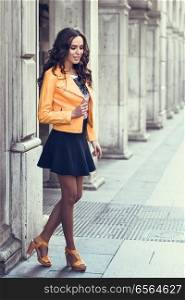 Young brunette woman, model of fashion, wearing orange modern jacket and blue skirt. Pretty caucasian girl with long wavy hairstyle. Female in urban background.