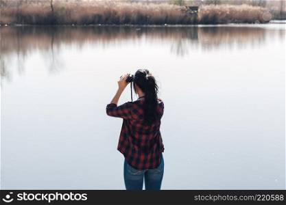 Young brunette woman on a lake&rsquo;s jetty with binoculars and plaid shirt