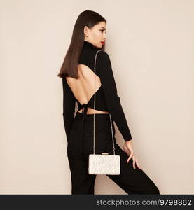 Young brunette woman posing in a black tight suit. Girl  with beige handbag. Brunette with long smooth and shiny hair. Fashion photo