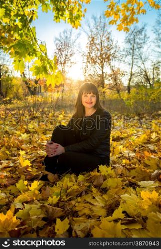 Young brunette woman sitting on grass at autumn park at sunny day