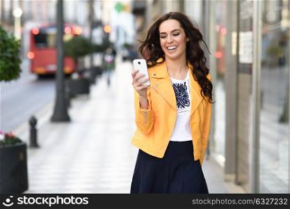 Young brunette woman smiling and looking at her smartphone, wearing orange modern jacket and blue skirt in urban background.