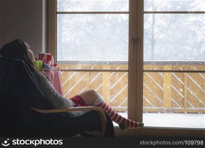 Young brunette woman wearing a cozy sweater and long red and white socks, sitting on an armchair in front of big glass door looking at the snowfall.