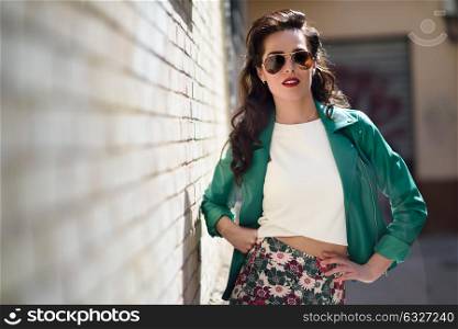 Young brunette woman with aviator sunglasses. Girl, model of fashion, wearing green modern jacket, standing in urban background.
